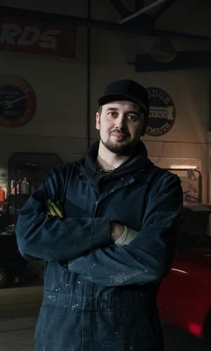 Experienced mechanic in a vintage garage standing confidently beside a classic car.